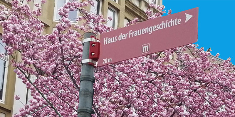 Straßenhinweisschild zum Haus der FrauenGeschichte vor dem Hintergrund eines Kirschblütenbaums